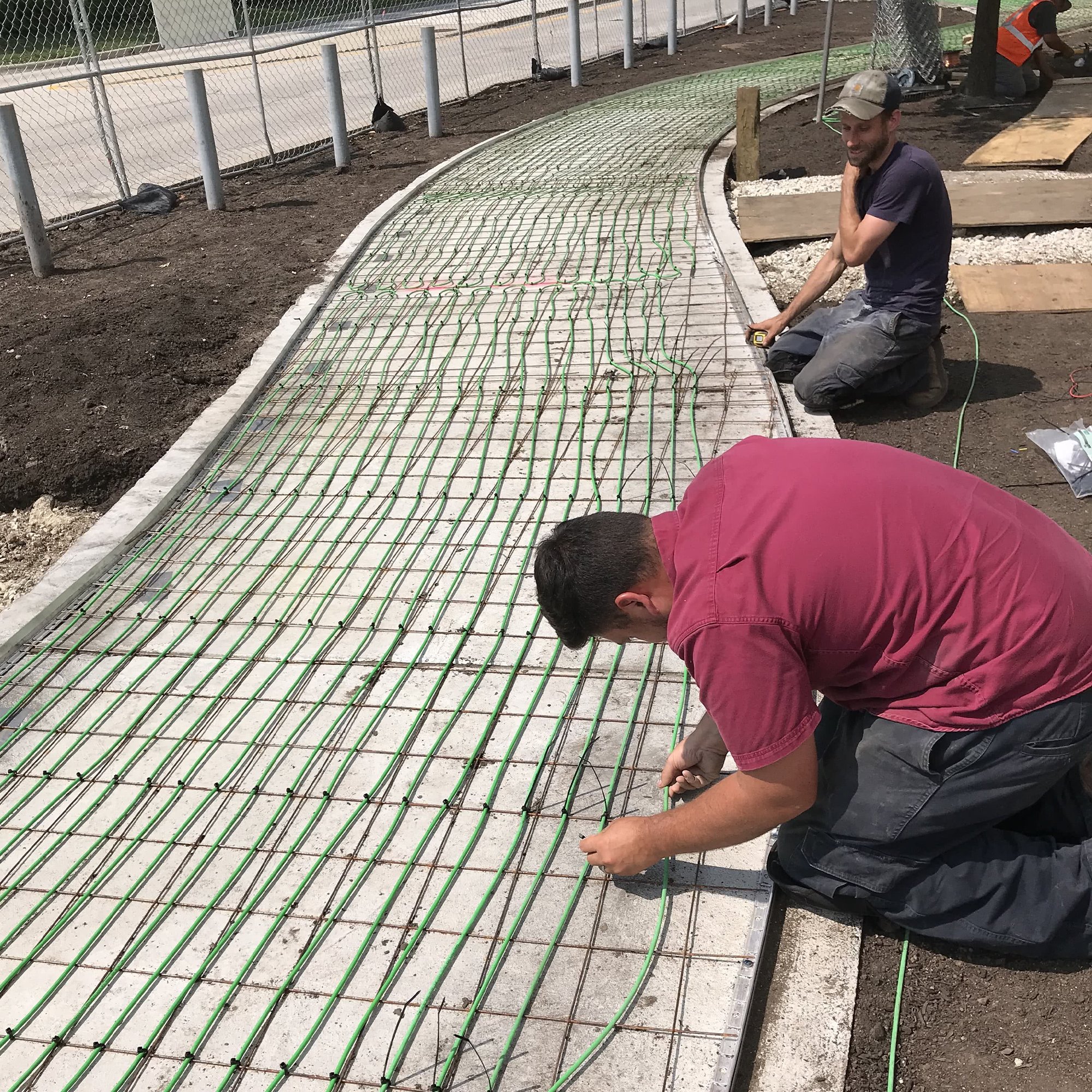 Workers securing green snow melting heating cable to wire mesh on a curved concrete walkway base.
