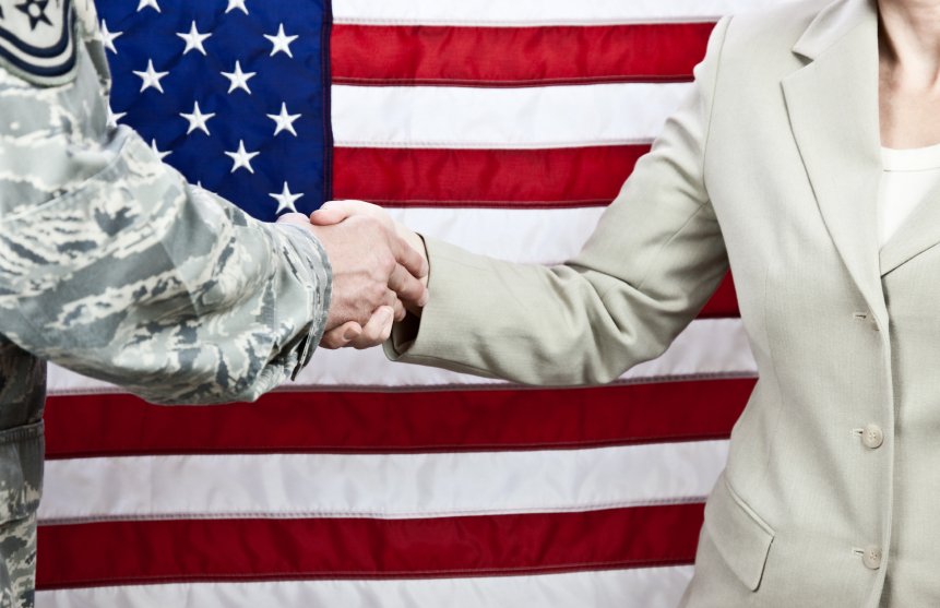 A person in a military uniform shaking hands with a person in a business suit in front of an American flag to honor veterans.