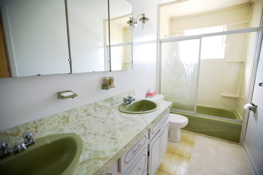 An outdated bathroom interior featuring an avocado-green bathtub, double vanity with green-veined countertops, and light-colored square floor tile.