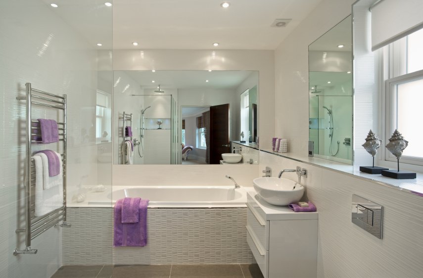 A polished metallic ladder-style towel warmer mounted on a white tiled bathroom wall holding white and purple towels.