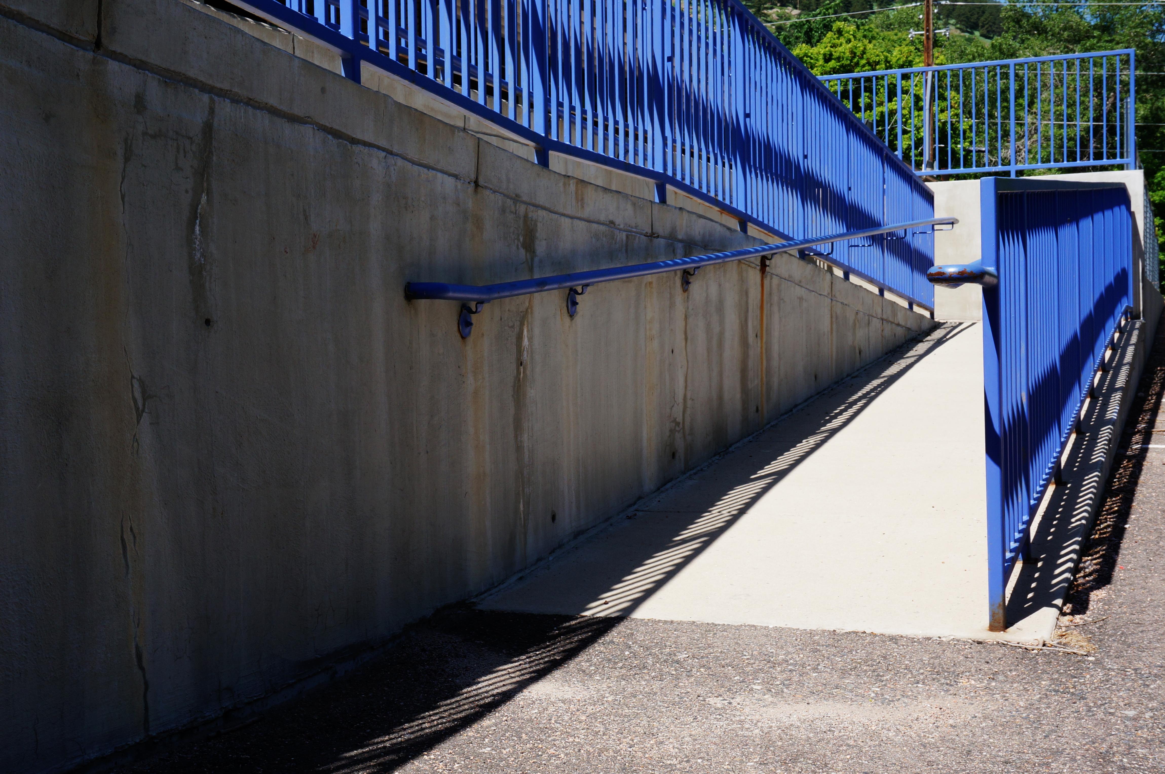A concrete accessibility ramp with blue metal railings rising alongside a concrete retaining wall in bright sunlight.