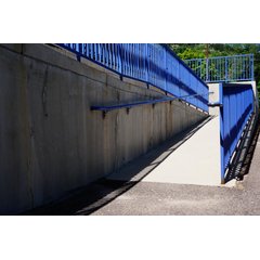 A concrete accessibility ramp with blue metal railings rising alongside a concrete retaining wall in bright sunlight.