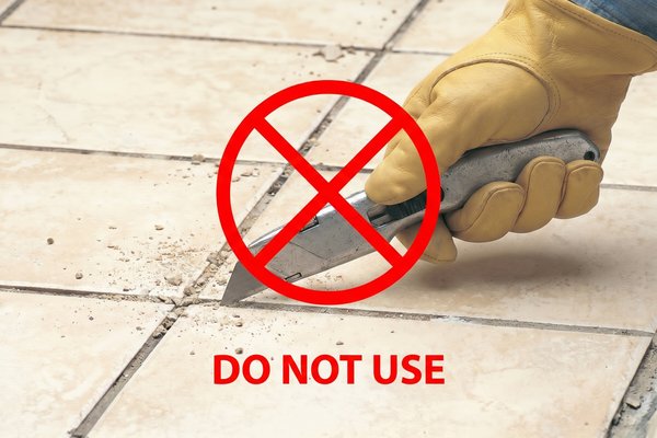 A gloved hand using a utility knife to scrape grout between installed floor tiles, marked with a prohibition symbol to warn against the practice.