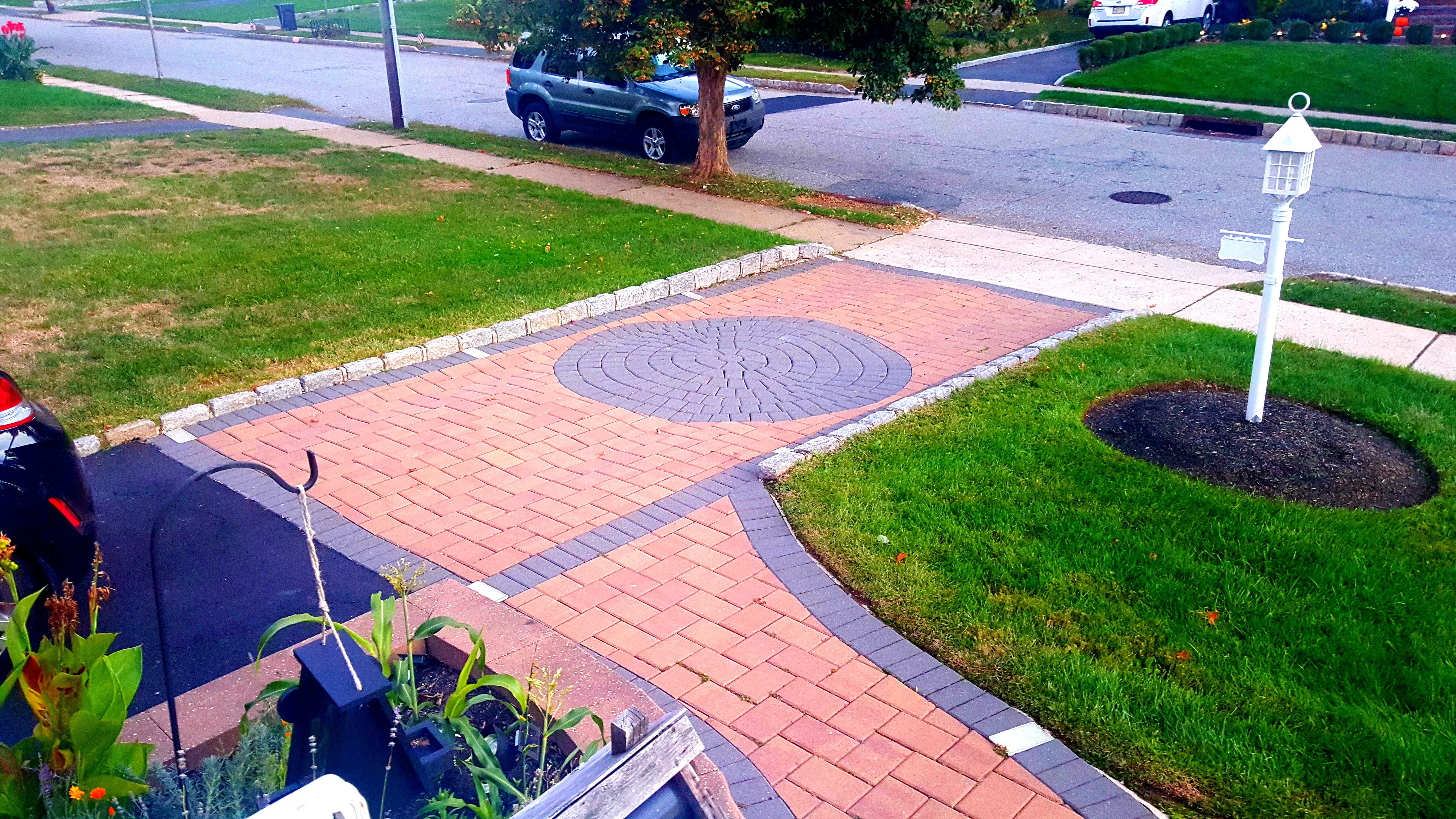 A residential driveway and walkway featuring interlocking reddish-tan and dark grey pavers with a central circular medallion.