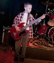 A young boy playing a red SG-style bass guitar on a stage with a drum kit and amplifier in the background.