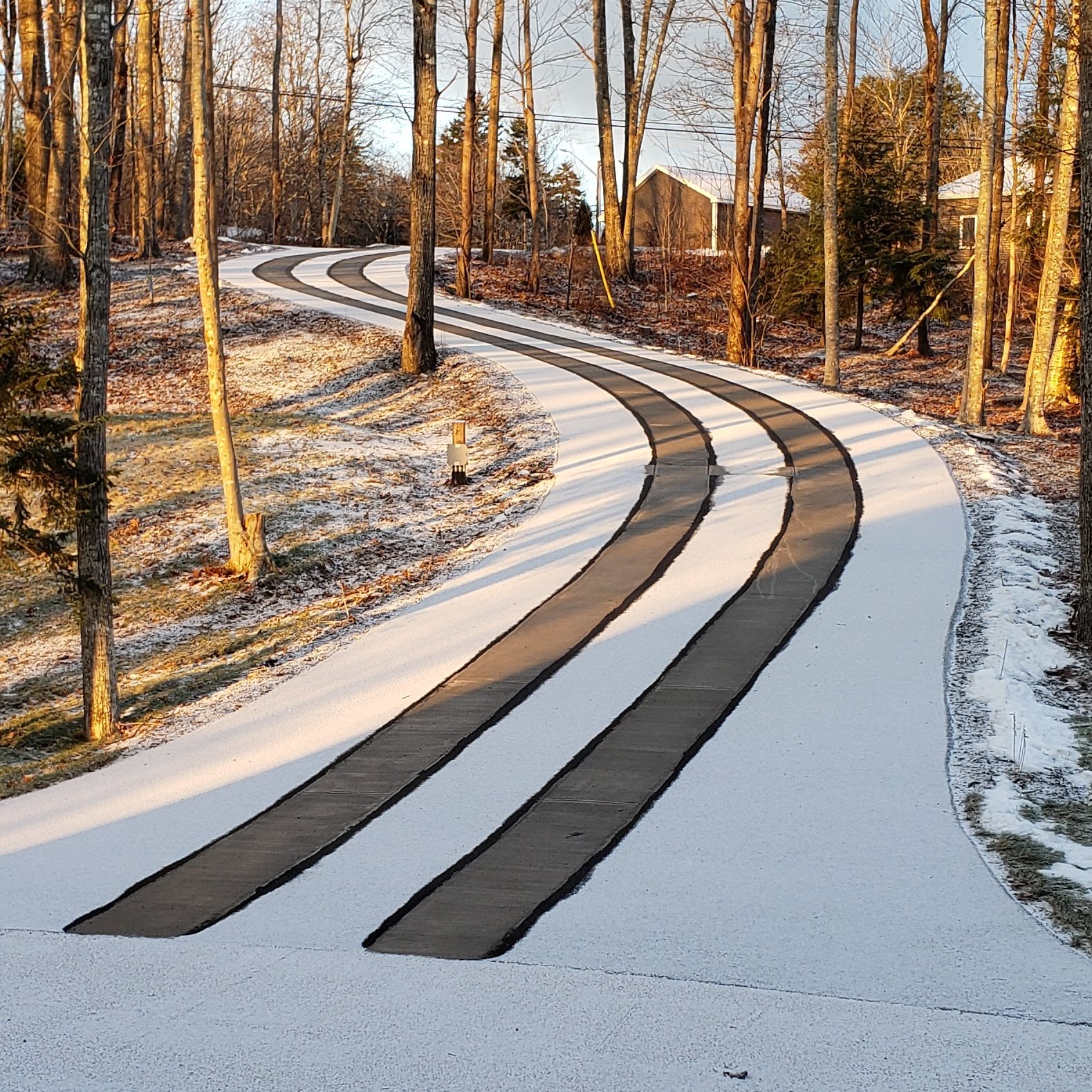 A snow melting system creates clear tire tracks on a snow-covered asphalt driveway, revealing the pavement beneath.