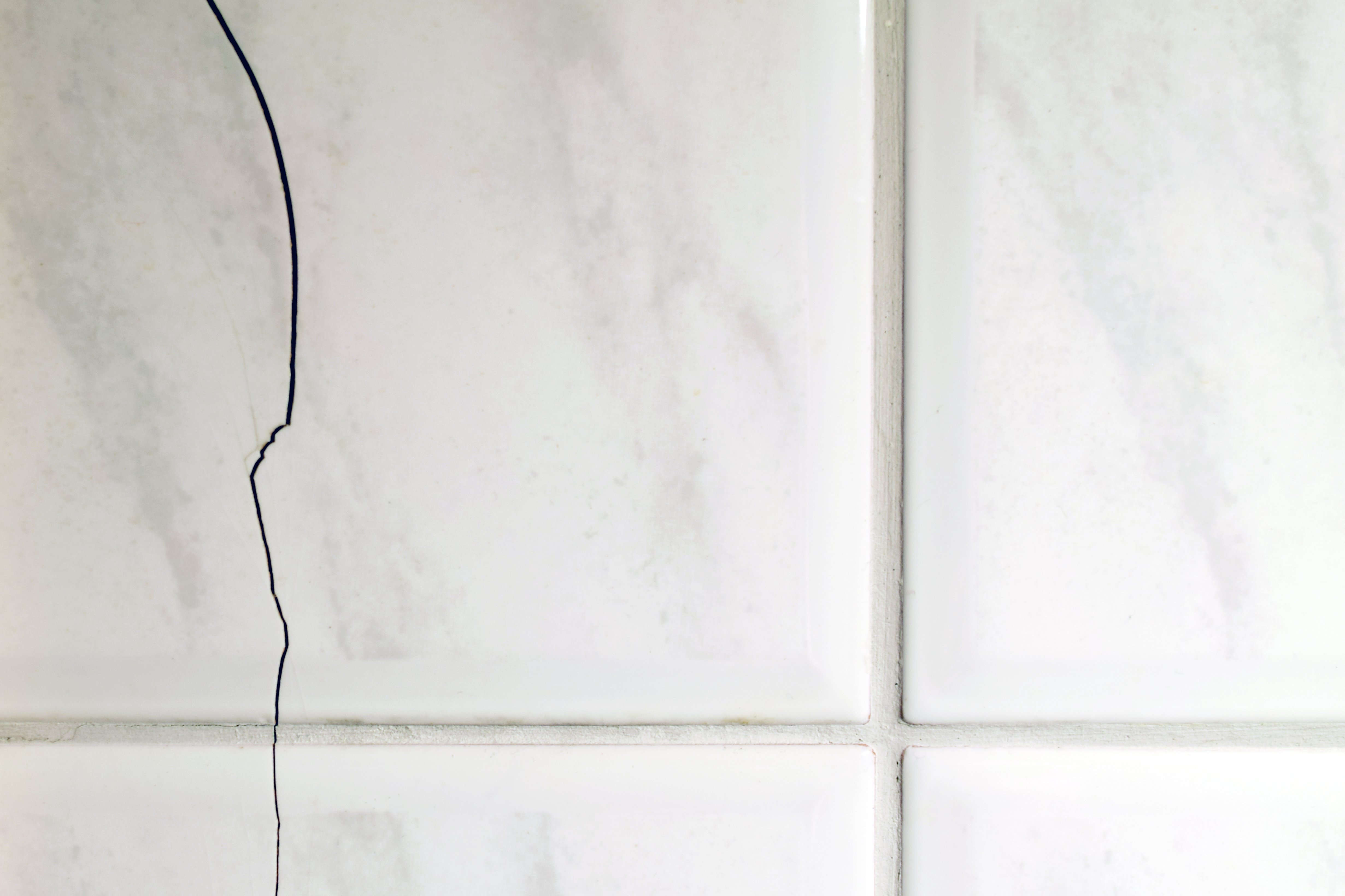A close-up of white marbled ceramic floor tiles featuring a prominent jagged crack extending across the tile surface and grout line.