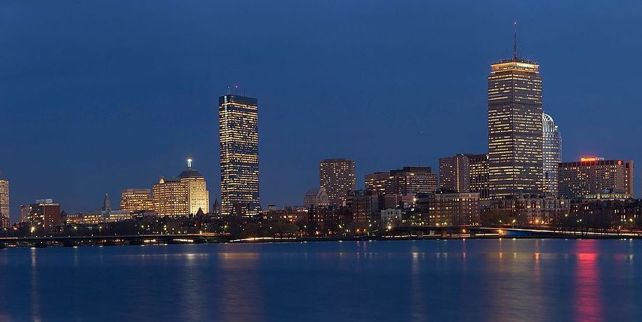 A nighttime panoramic view of the illuminated Boston skyline reflected in the Charles River at dusk.