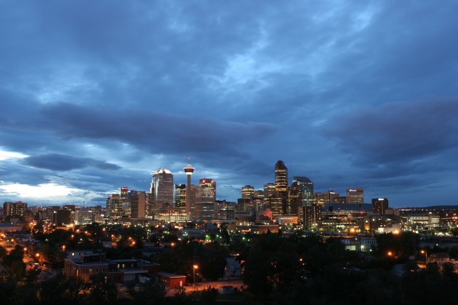The Calgary city skyline at dusk with illuminated skyscrapers and the Calgary Tower glowing under a dramatic cloudy blue sky.