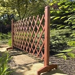 A freestanding expandable wooden trellis fence with a diamond lattice pattern installed on a stone path in a garden.