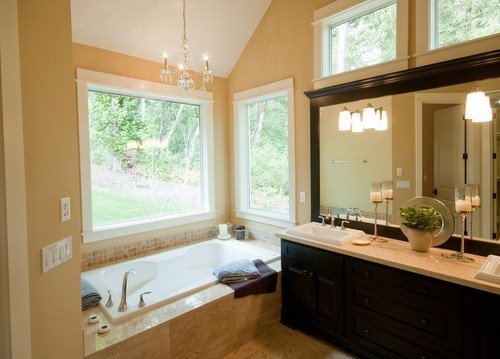 A bright, comfortable bathroom featuring a drop-in bathtub surrounded by stone tile and a dark wood vanity with a stone countertop.