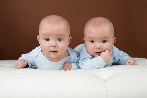Twin baby boys in blue onesies lying on a white tufted surface in a bedroom setting.