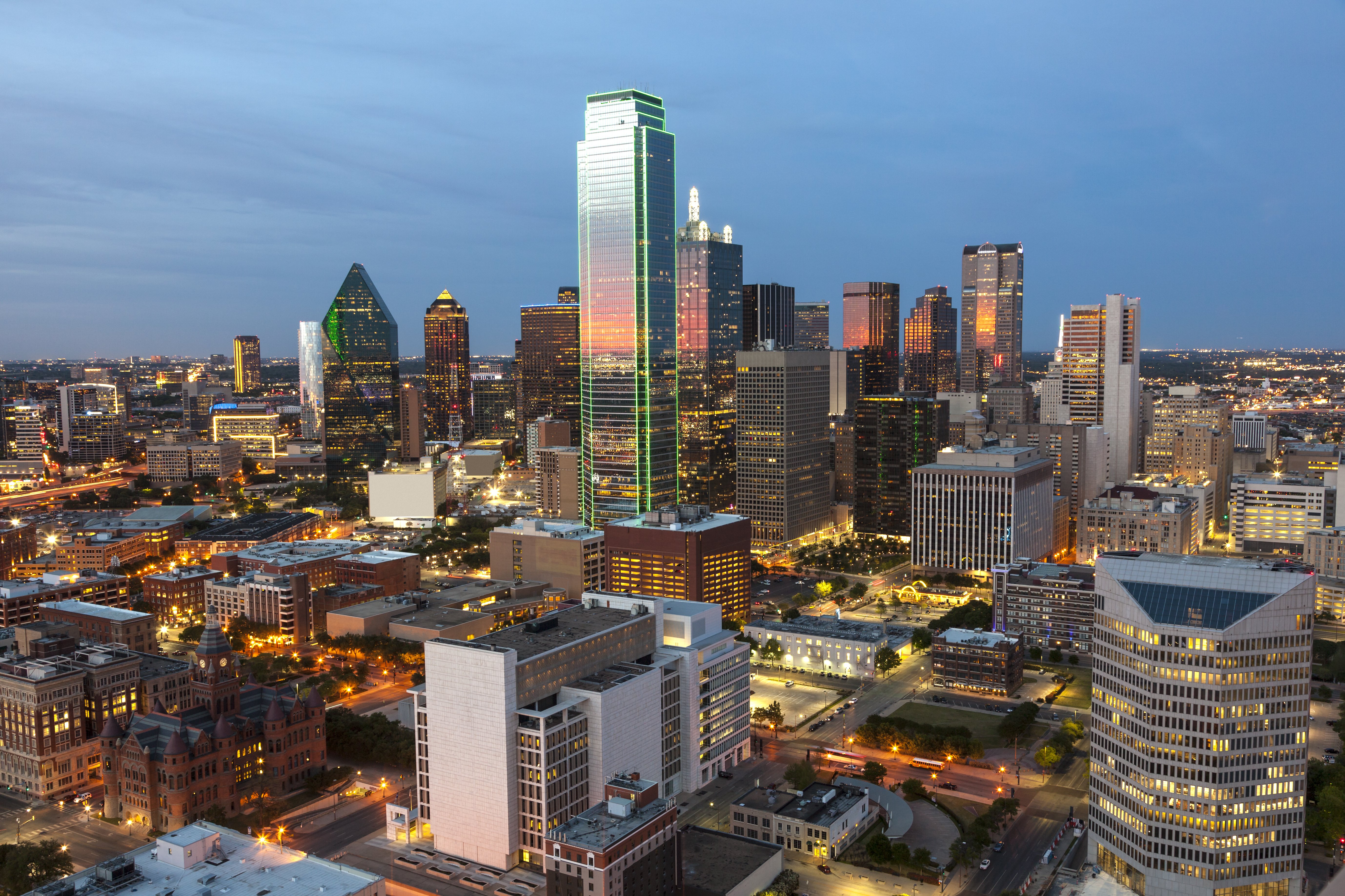 Aerial view of the Dallas skyline at dusk featuring the Bank of America Plaza skyscraper illuminated with green neon lighting.