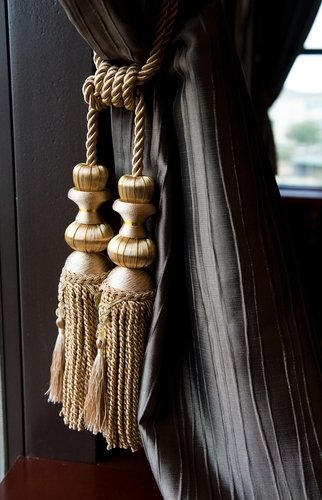 Decorative dark taupe fabric curtain held back by a thick twisted rope tieback featuring ornate gold-toned silk-wrapped tassels.