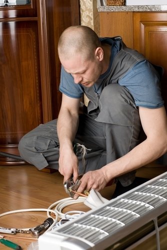 A technician in work overalls using a wrench to tighten a brass fitting on copper piping for an indoor mini-split air conditioning unit.