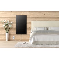 A black glass Ember infrared heating panel mounted vertically on a light-wood plank wall in a bedroom.