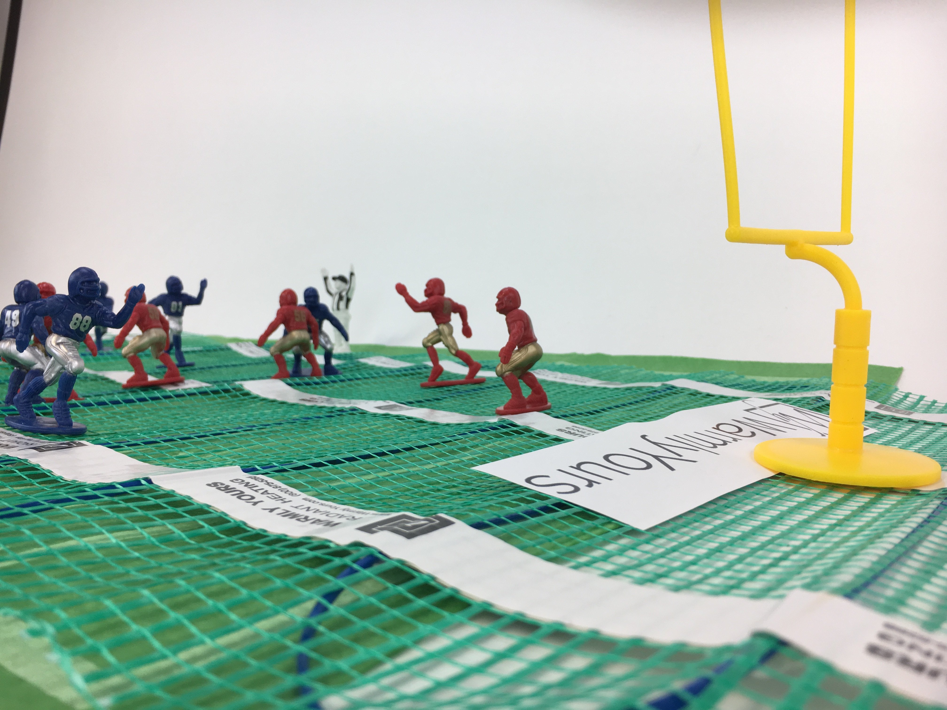 Miniature football figures arranged on a WarmlyYours TempZone electric radiant floor heating mat with blue heating cable on green mesh.