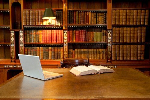 A traditional home office featuring a large wooden desk with an open laptop and book in front of an expansive, well-lit library bookcase.