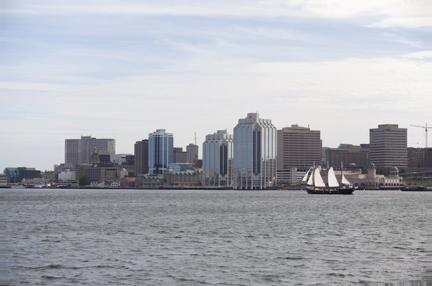 A wide view of the Halifax NS skyline featuring modern office buildings and a sailing ship on the water under a partly cloudy sky.