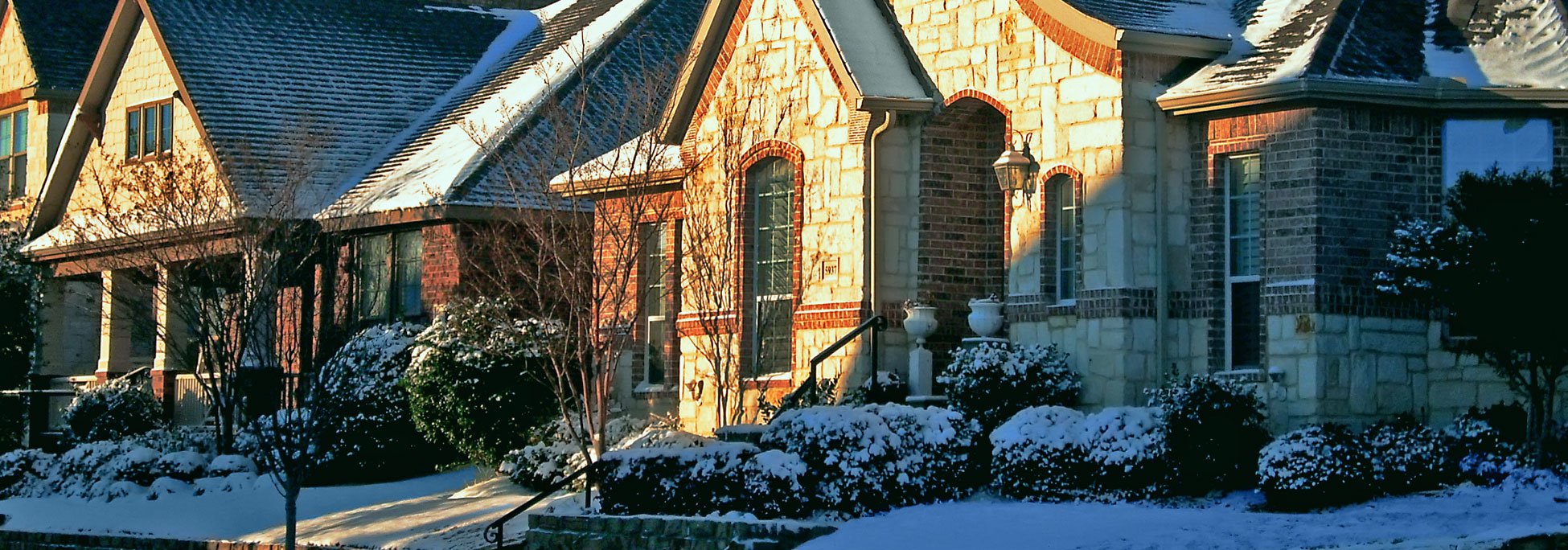 A snow-covered residential neighborhood in winter with sunlight illuminating the stone facade of a home.