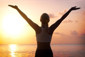 A woman in athletic attire standing by calm water at sunrise, reflecting the healing effects of radiant heat.
