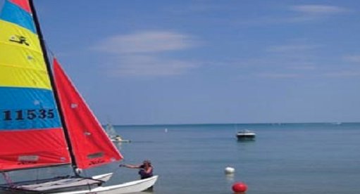 A small catamaran sailboat with colorful sails anchored in the calm waters of the Highland Park lakefront.