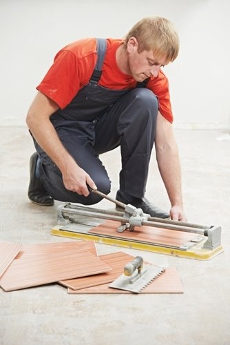 A professional tiler kneeling on a subfloor while using a manual tile cutter to trim a reddish-brown ceramic tile.