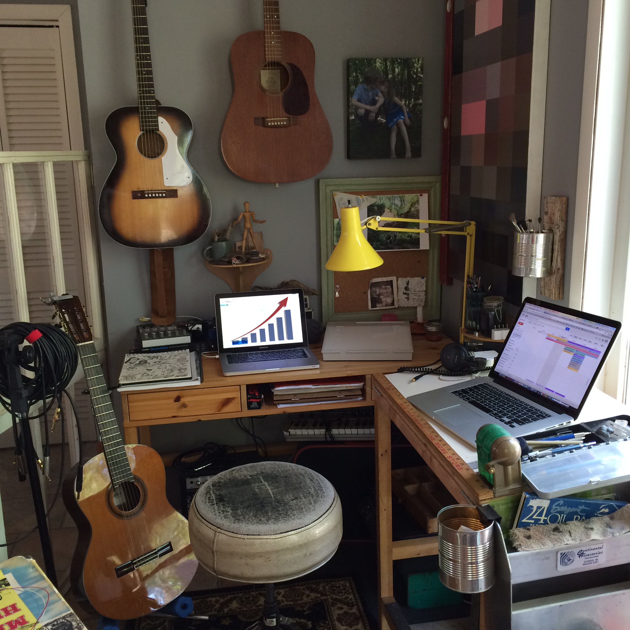 A home office and music studio corner featuring acoustic guitars on the wall, a wooden desk with two laptops, and a yellow desk lamp.