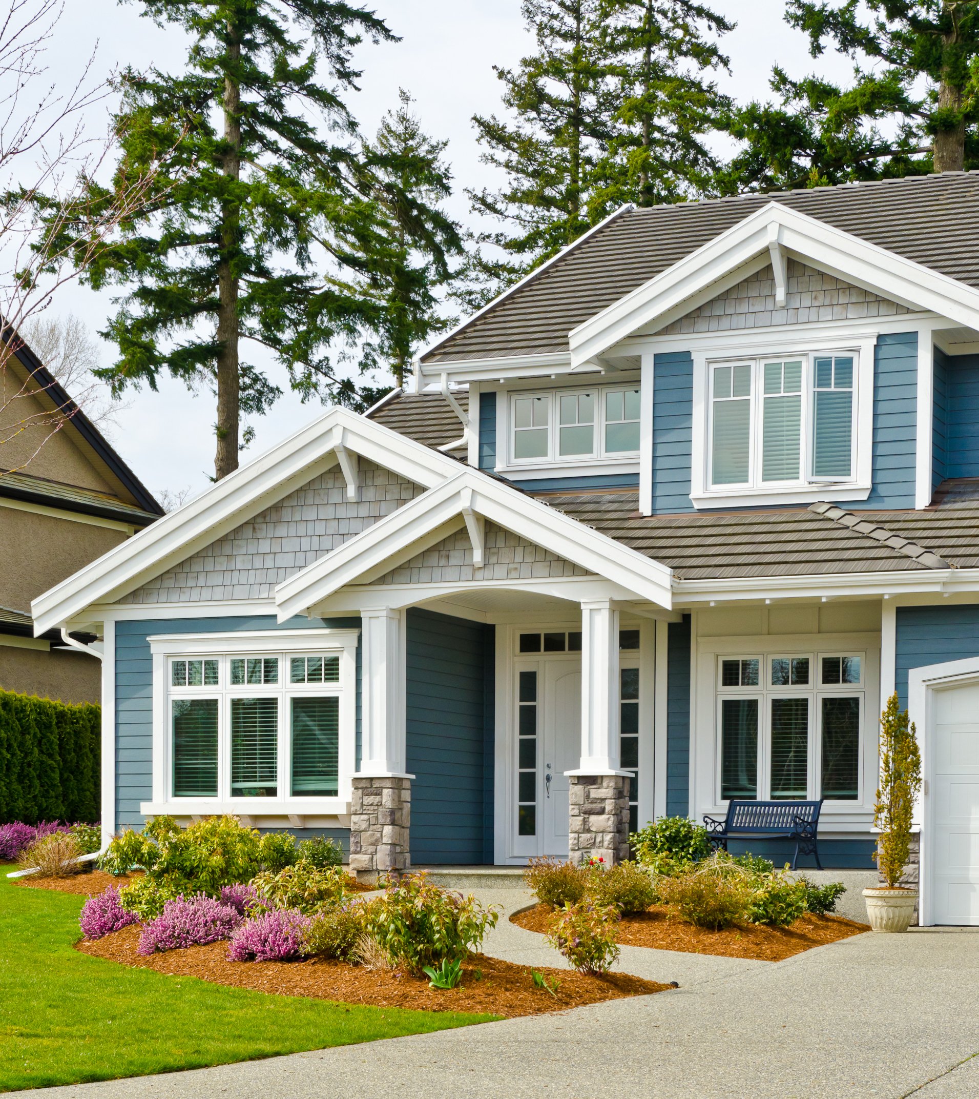 A two-story craftsman-style house with blue and grey siding, featuring an aggregate concrete walkway and driveway.