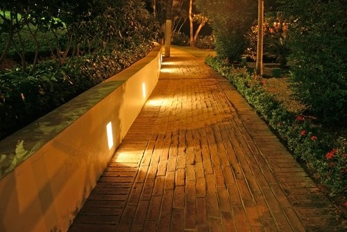 A nighttime view of a curved brick paved walkway illuminated by recessed wall lights and surrounded by lush landscaping.