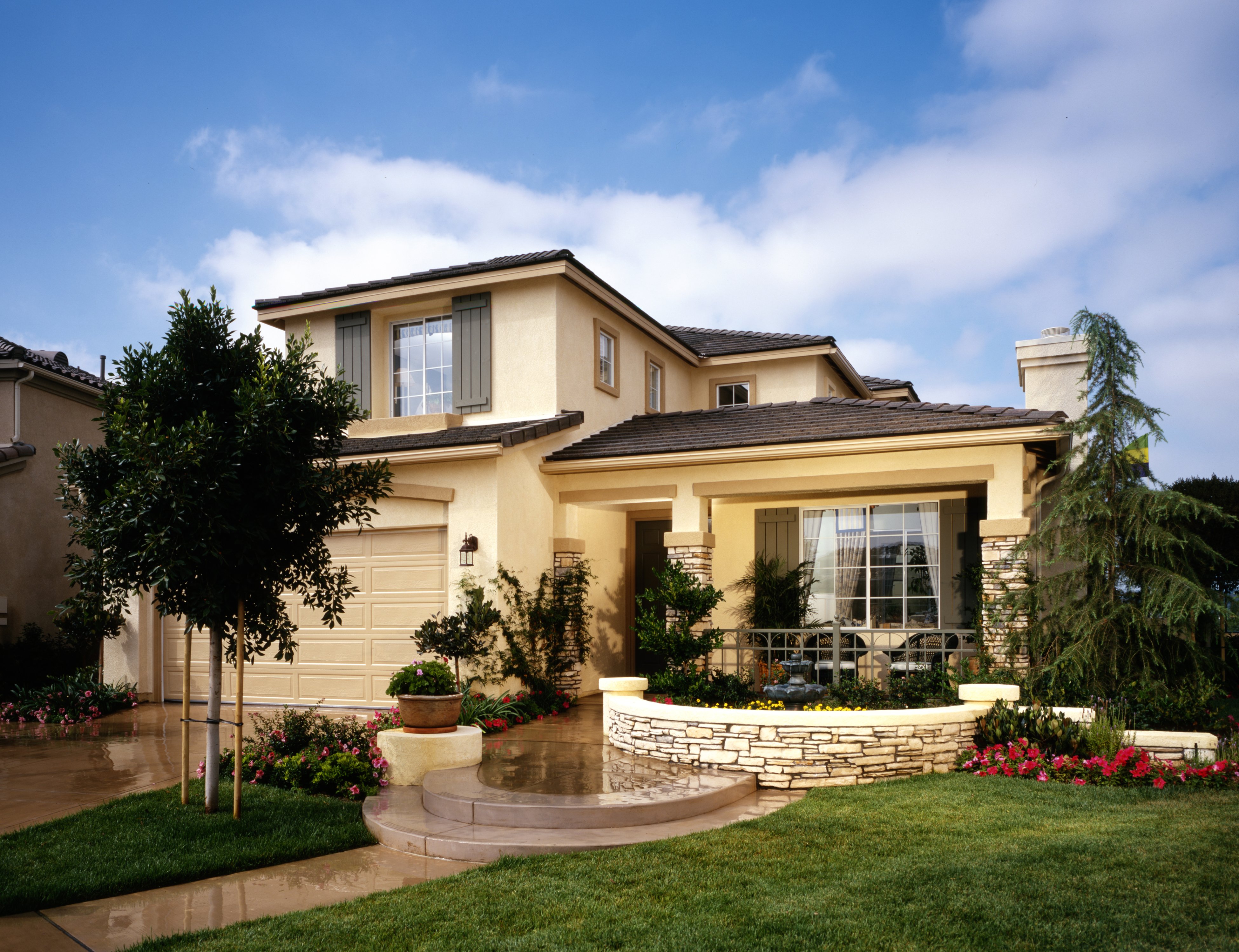 A two-story beige stucco house with a landscaped front yard, stone-accented porch, and a concrete walkway leading to a curved stone fountain.