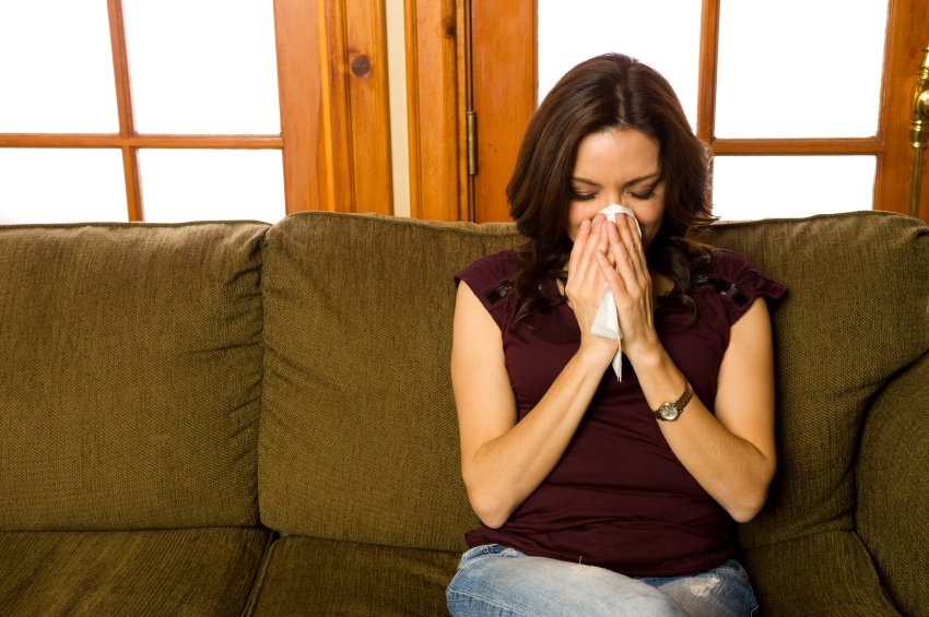 A woman sitting on an olive-green sofa holding a tissue to her nose in a home interior.