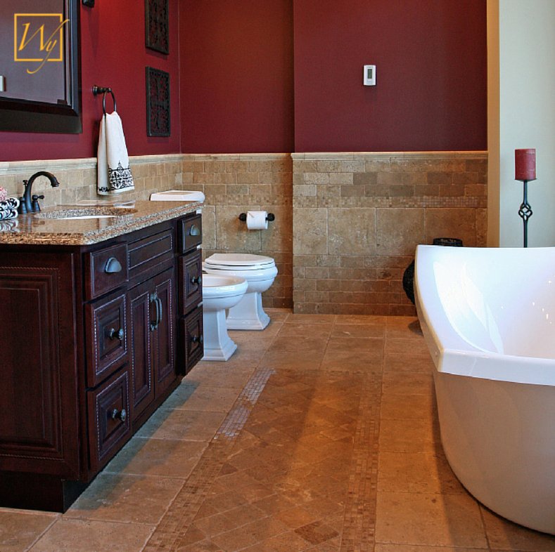 A traditional bathroom with natural stone tile flooring featuring an electric radiant floor heating thermostat mounted on the burgundy wall.