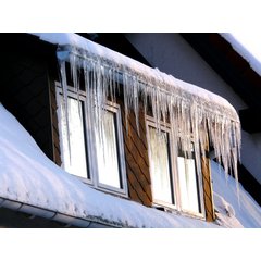 A snow-covered house dormer with long icicles hanging from the roof edge above four window panes.