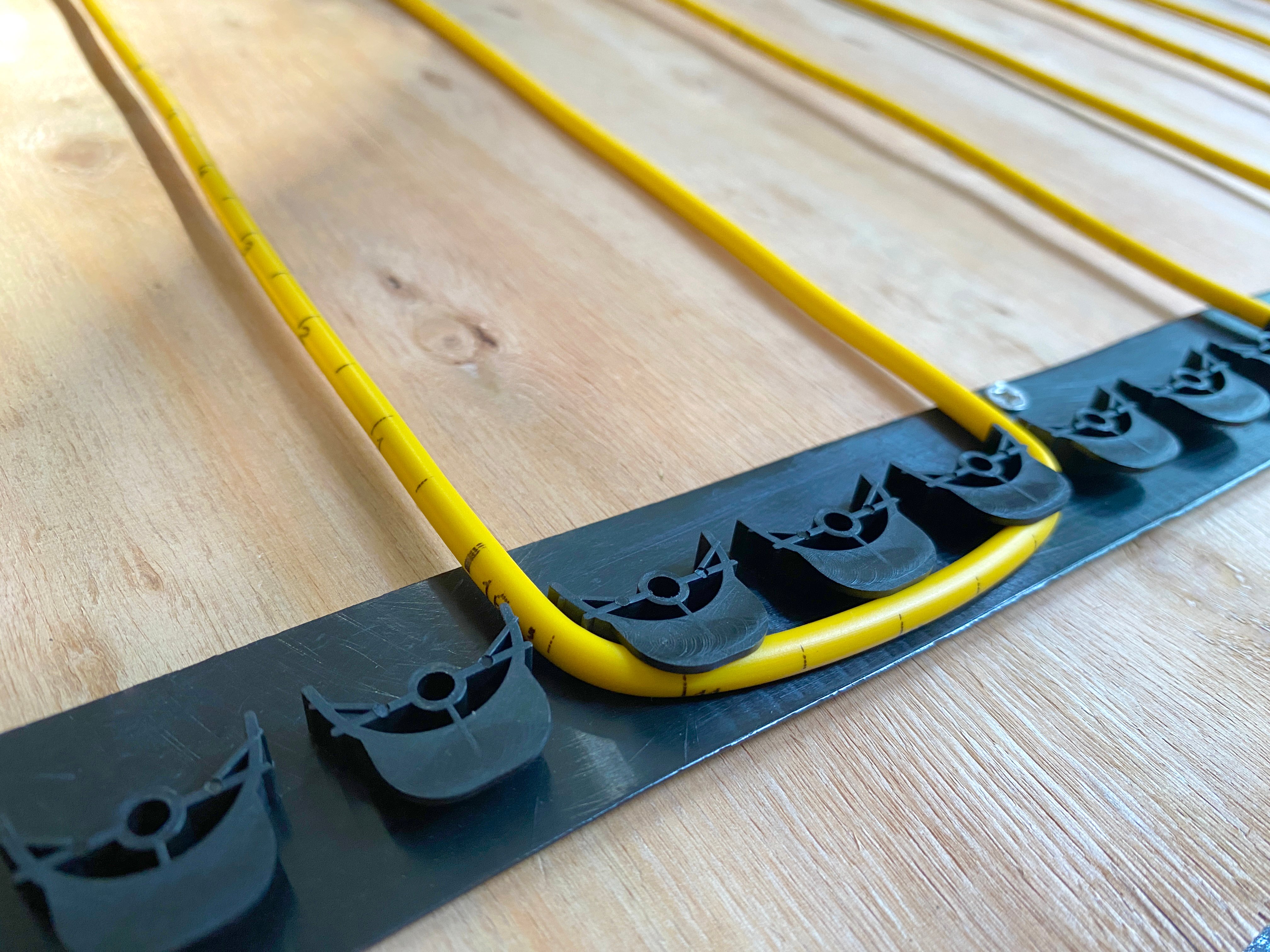 Yellow TempZone Ruler Cable with measurement markings installed in parallel loops on a wood subfloor using black GripStrip fixing strips.