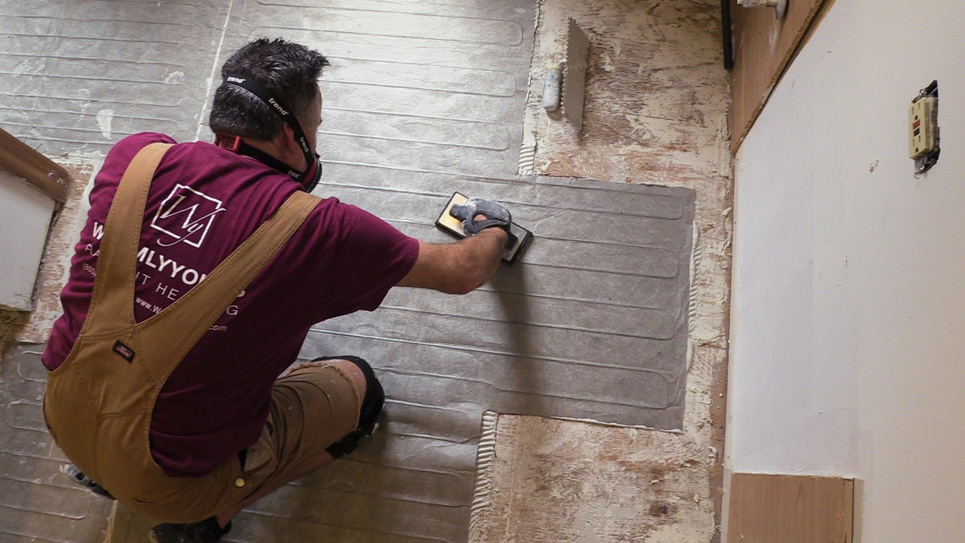 An installer embeds a TempZone Custom Mat electric radiant floor heating mat into thin-set mortar over a plywood subfloor.