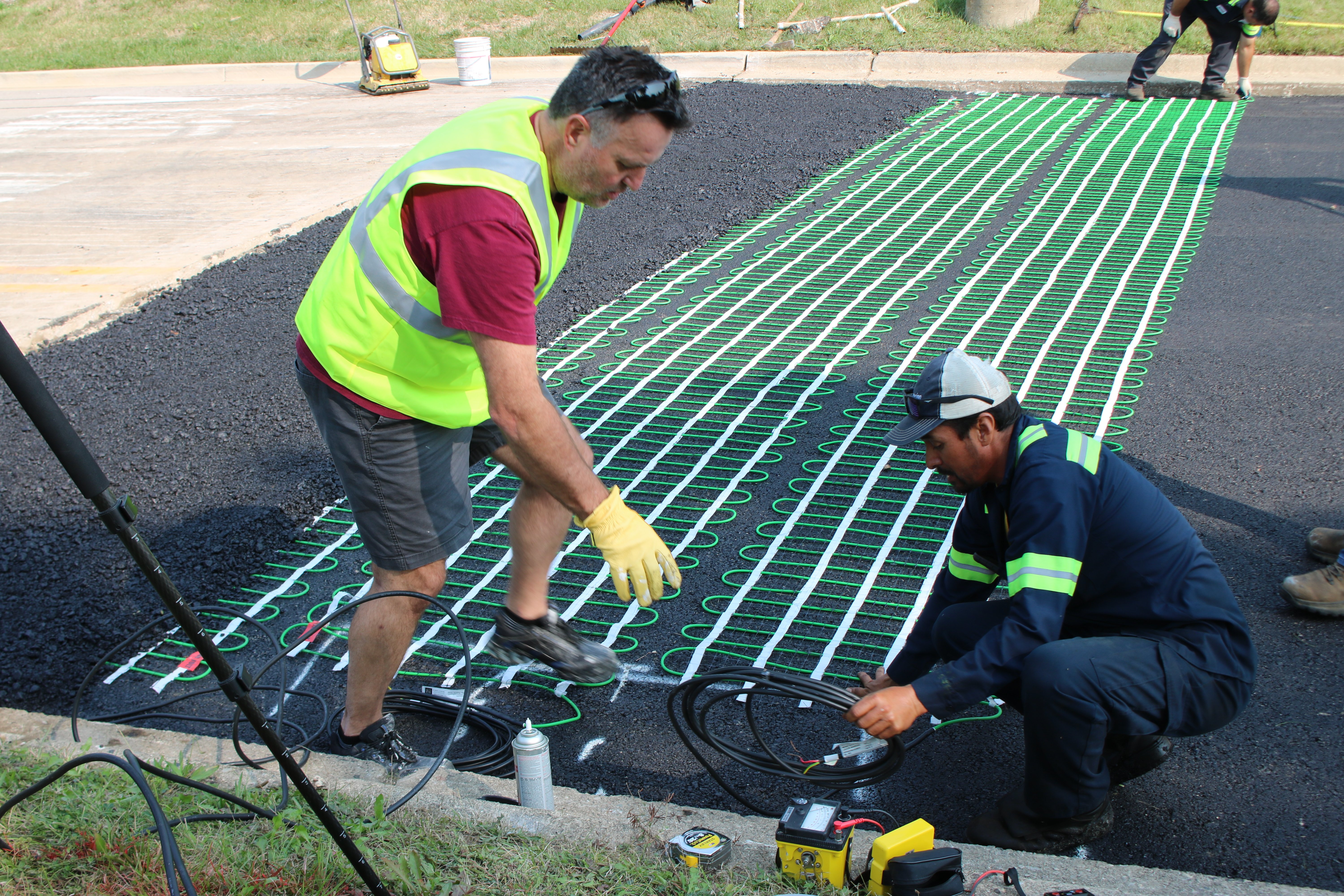 Workers installing a snow melting heating mat with green heating cable on white mesh backing onto an asphalt base.