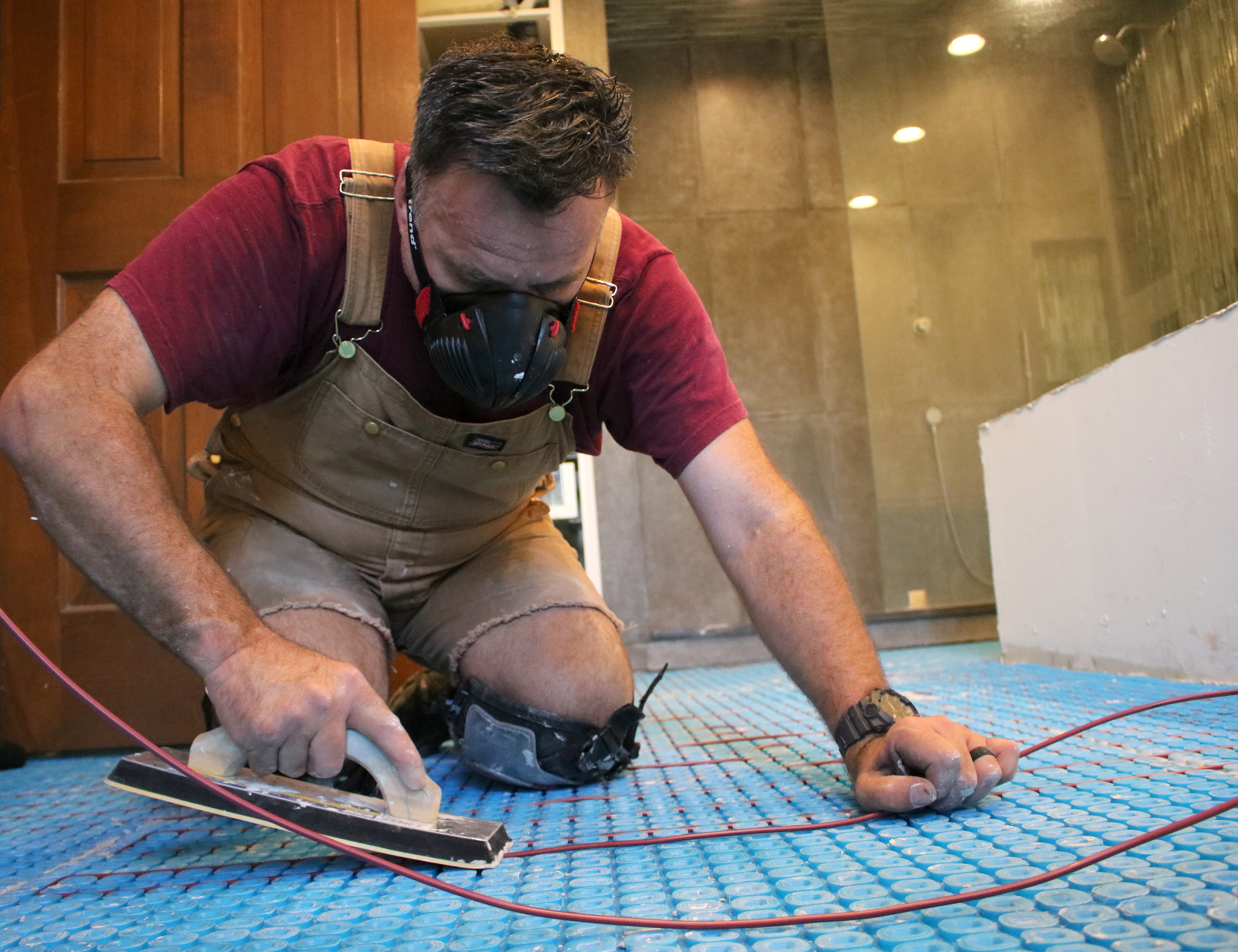 An installer using a rubber float to press TempZone electric radiant floor heating cable into a blue uncoupling membrane in a bathroom.