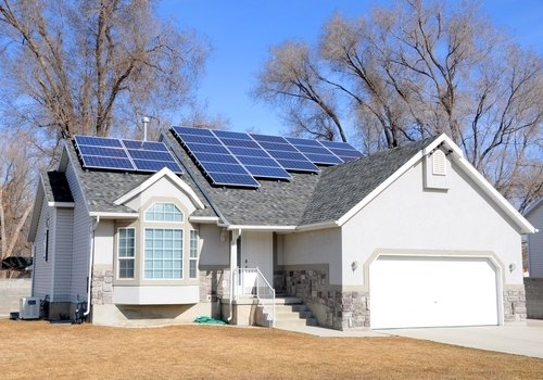 A suburban single-story house with solar panels installed on the roof under a clear blue sky.