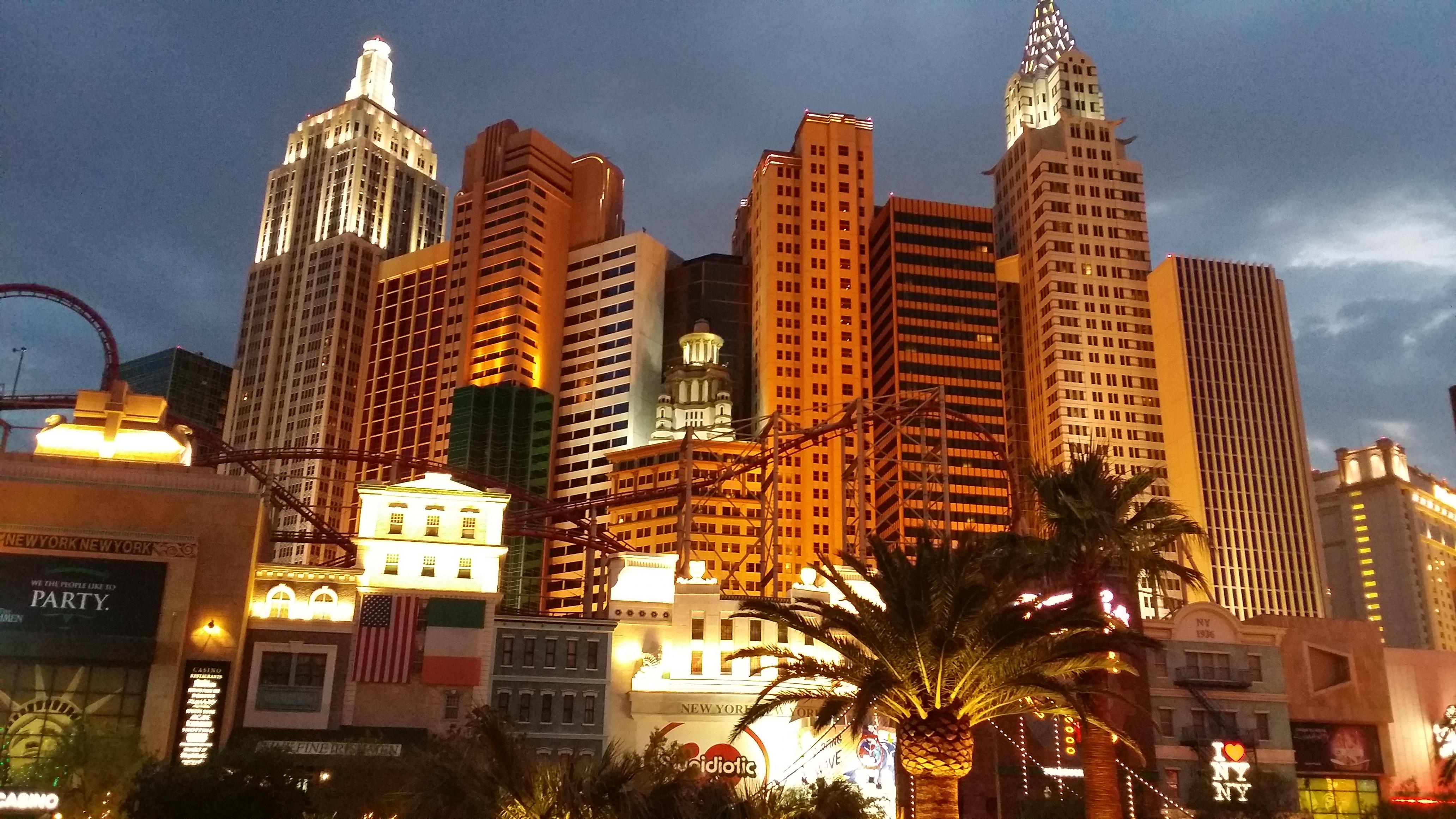 The illuminated New York-New York Hotel and Casino facade on the Las Vegas Strip at dusk featuring skyscraper replicas and roller coaster tracks.
