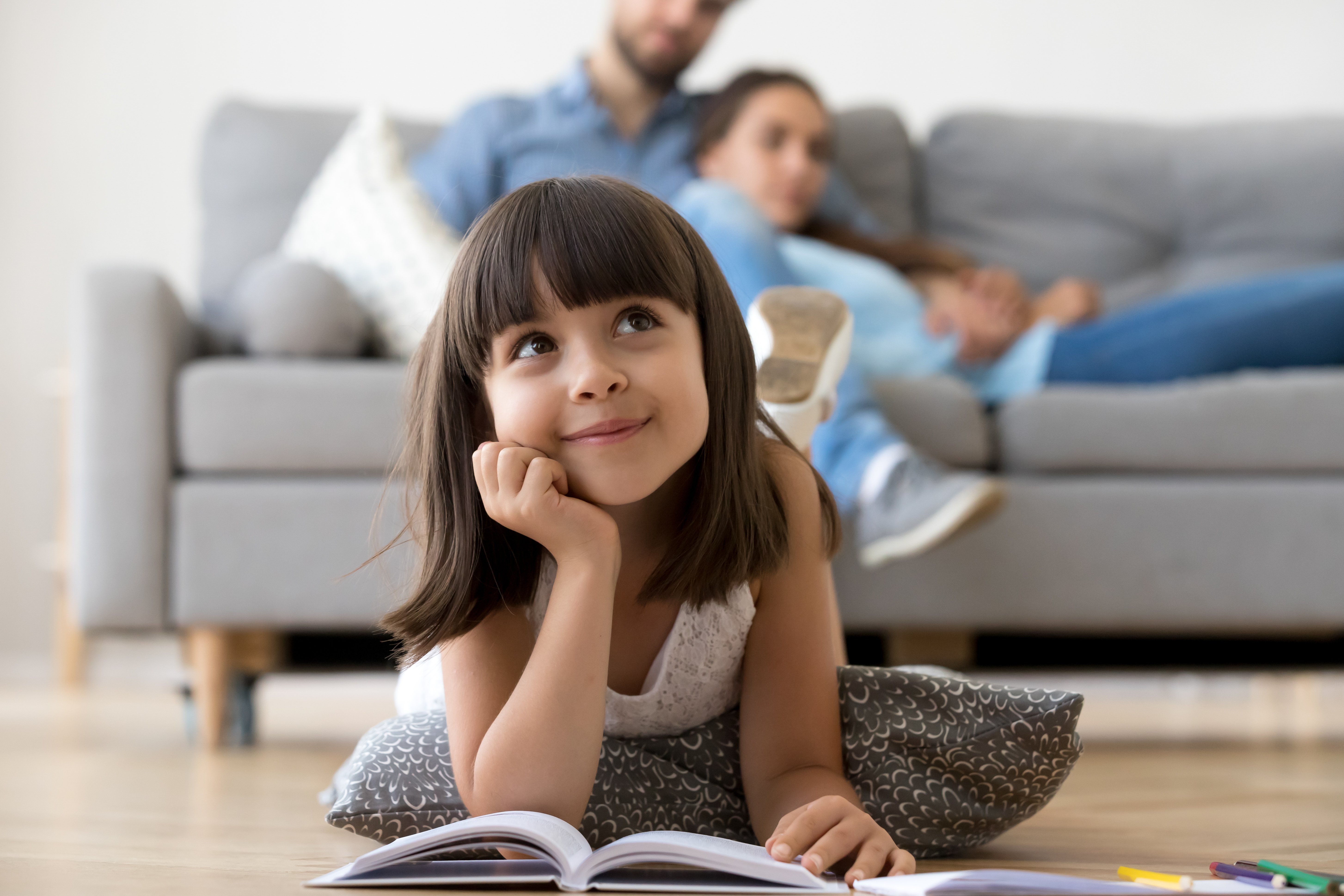 A young girl reading a book while lying on a wooden floor in a living room.