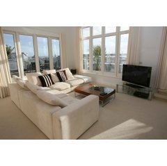 A bright living room interior featuring a cream sectional sofa and glass coffee table with a view of an outdoor balcony and harbor.