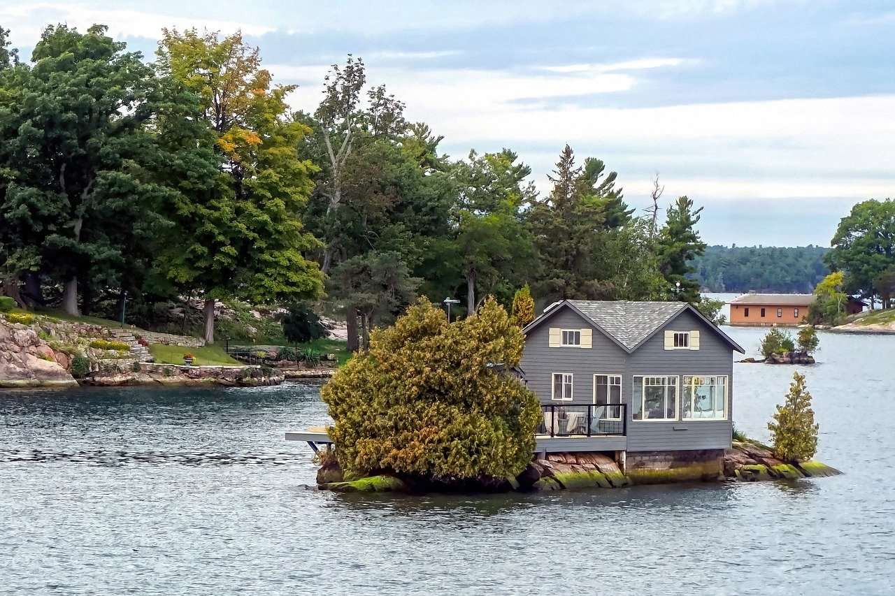 A grey cottage with double-gabled siding and a glass-enclosed porch situated on a small rocky island surrounded by water.
