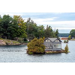 A grey cottage with double-gabled siding and a glass-enclosed porch situated on a small rocky island surrounded by water.