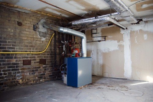 An unfinished basement utility room featuring a blue and grey boiler, exposed brick wall, and overhead insulated piping.