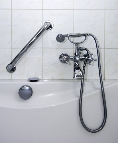 A vintage-style chrome bathtub faucet set with a handheld shower head mounted on a wall of pale marble-patterned ceramic tiles.