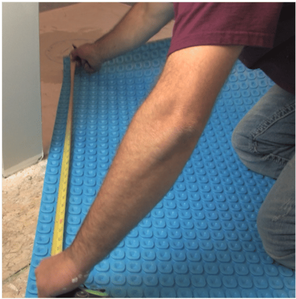 A person measuring a blue Prodeso uncoupling membrane installed on a subfloor for an electric radiant floor heating system.
