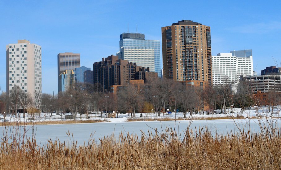 A winter view of the Minneapolis skyline featuring snow-covered parkland and high-rise buildings across a frozen lake.