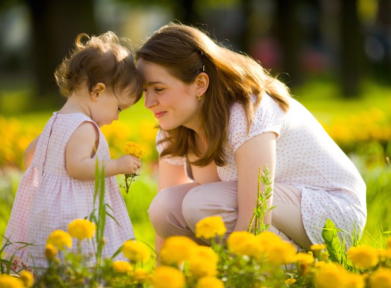 A mother and toddler interacting in a sunny garden with yellow flowers in the foreground.