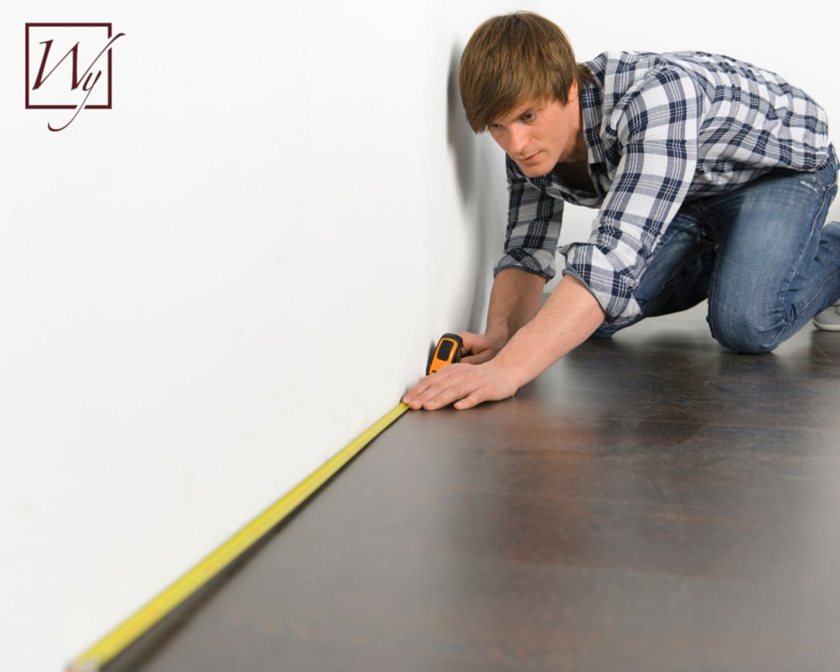 A man measuring a room with a tape measure along the base of a wall for a WarmlyYours radiant heating installation.